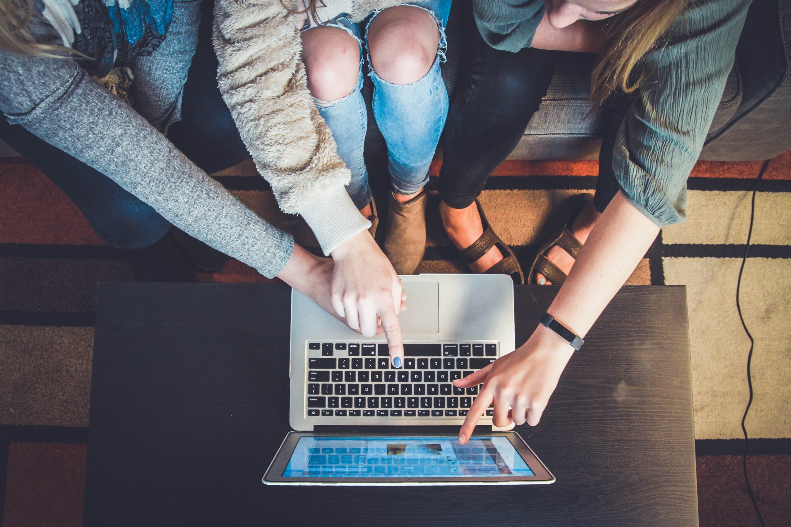 a number of people surrounding a laptop on a wooden table, one person pointing at the laptop