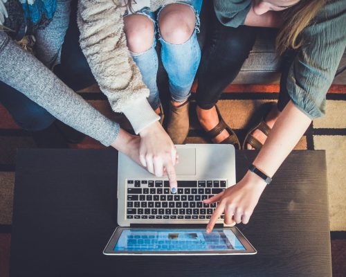 a number of people surrounding a laptop on a wooden table, one person pointing at the laptop